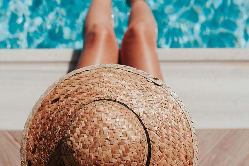 Woman sat with her feet in a pool wearing a straw hat for a sun skin care step to protect her hair from the sun